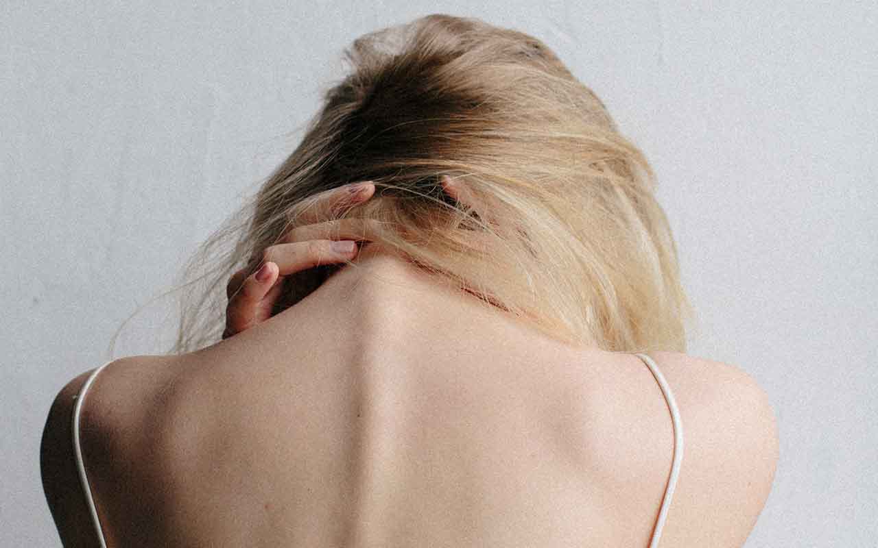 mujer rubia de espalda se sujeta la nuca agachando la cabeza, se ve su espalda cervical y dorsal lleva unos tirantes blancos, el fondo es gris
