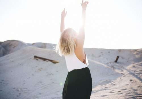 Mujer de espalda eleva los brazos hacia el cieloy la mirada con el sol justo delante del rostro muy luminoso esta feliz dentro de unas dunas de arena