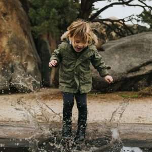 Niño de aproximadamente 3 años vestido de zapatos negros, pantalon negro, chaqueta verde saltando dentro de un charco de agua, con cara sonriente, detras de el se observa sueo de arean, piedras y un arbol