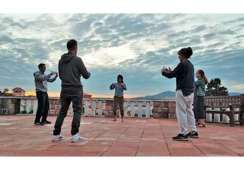 se ve un grupo de 5 personas de pie en circulo practicando Qi Gong en una terraza del parque de Montjuic en Barcelona, en el fondo se ve el cielo luminoso del atardecer